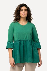 Das grüne Baumwolloberteil hat einen V-Ausschnitt, dreiviertellange Ärmel mit Rüschenrand und eine geraffte Taille für einen schichtenähnlichen Effekt.