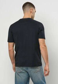 Mann med kort hår iført en enkel svart t-skjorte og blå jeans, stående vendt mot en lys grå vegg.