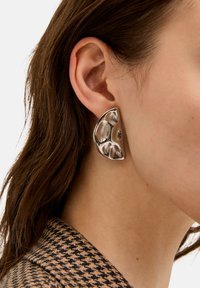 Primo piano di un orecchio femminile che indossa un grande orecchino in argento di forma astratta, con capelli castani e un colletto di giacca a scacchi marrone e nero.