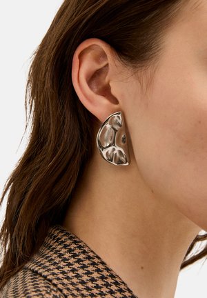 Primer plano de la oreja de una mujer que lleva un gran pendiente de plata con forma abstracta, con cabello castaño y un cuello de chaqueta de tejido houndstooth en tonos marrón y negro.