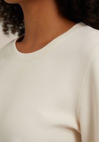 Svetlo béžová mikina s okrúhlym výstrihom, mäkkou textúrou a vsadenými rukávmi. Obsahuje jemné šitie a hladký materiál.