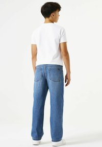Jean en denim bleu à coupe décontractée, doté de poches arrière et d'une fermeture à bouton. Porté avec un t-shirt blanc uni et des baskets blanches.