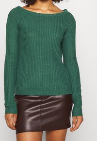 Grüner gestrickter Pullover mit langen Ärmeln, weitem Ausschnitt und gerippten Bündchen, kombiniert mit einem braunen Minirock aus Kunstleder mit glatter Oberfläche.