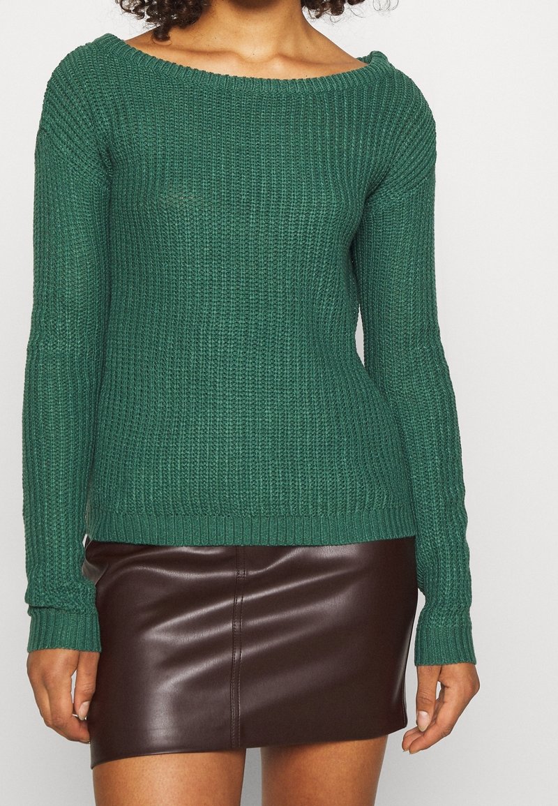 Grüner gestrickter Pullover mit langen Ärmeln, weitem Ausschnitt und gerippten Bündchen, kombiniert mit einem braunen Minirock aus Kunstleder mit glatter Oberfläche.