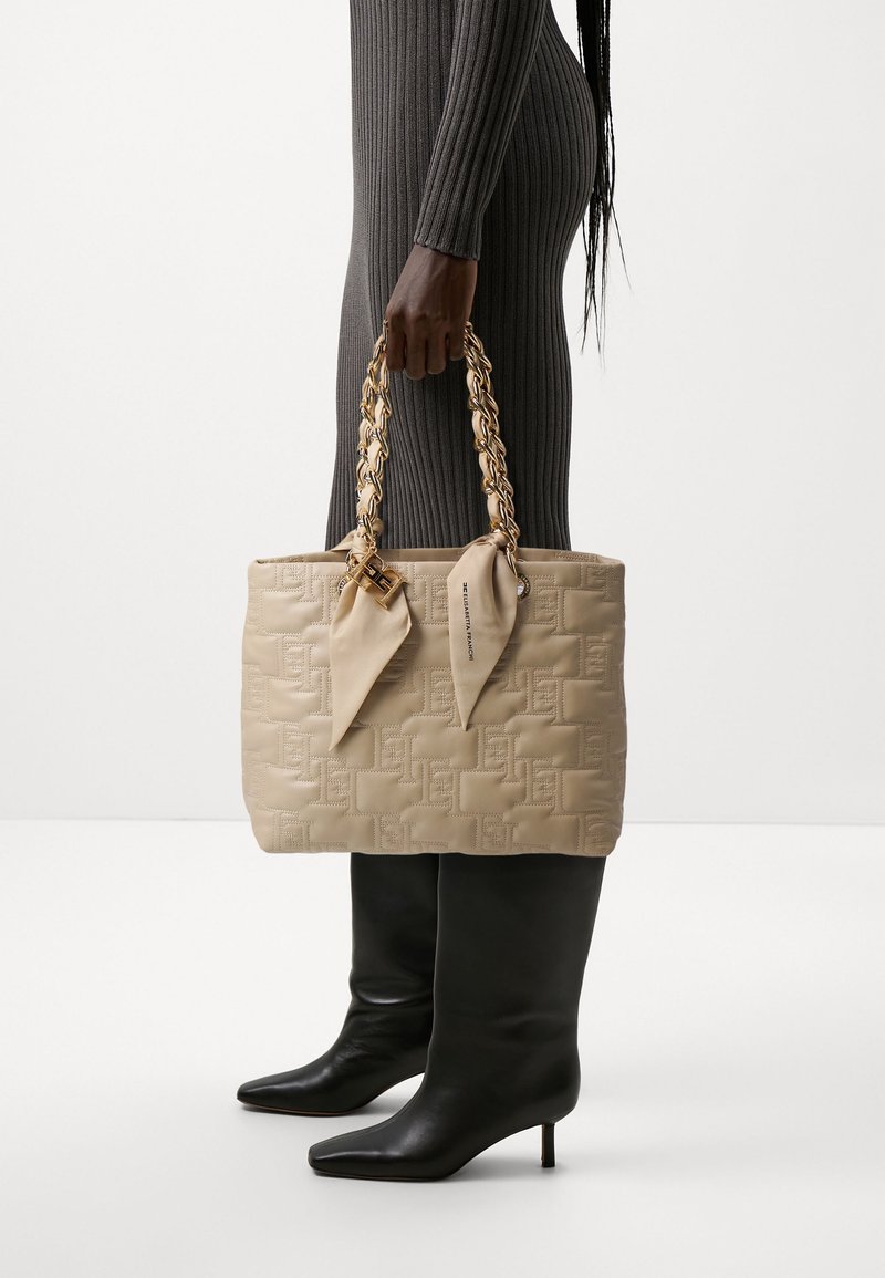 Personne en robe grise côtelée et bottines noires à talons tenant un sac à main matelassé beige avec des poignées en chaîne dorée et des nœuds en tissu.