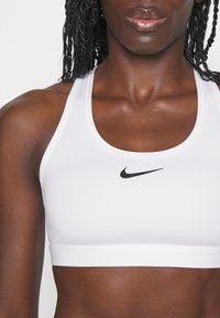 Bela športna modra iz raztegljive tkanine s ploskim pasom. Ima črn Nike logotip na sredini in zasnovo z zaobljenim izrezom.