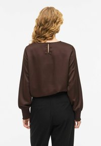 Brūna satīna blūze ar garām, pūstošām piedurknēm, aizmugurē ar atvērumu atslēgas formā un teksturētām elastīgām aprocēm, kopā ar gludiem melniem biksēm.