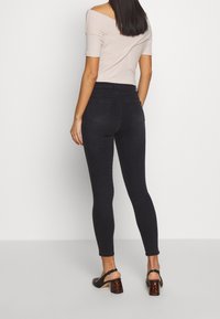 Jeans ajustados negros de cintura alta con bolsillos traseros estándar, combinados con una blusa clara de hombros al descubierto y sandalias negras de tacón.