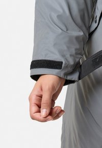 Grijze buitenkleding met een gladde textuur, voorzien van een klittenbandsluiting op de mouw en een licht losse fit, getoond met een hand die de mouw vastgrijpt.