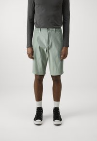 Pantalones cortos de color verde claro hechos de algodón, con un diseño clásico y cierre de botón, combinados con una camisa de manga larga oscura y zapatillas negras.