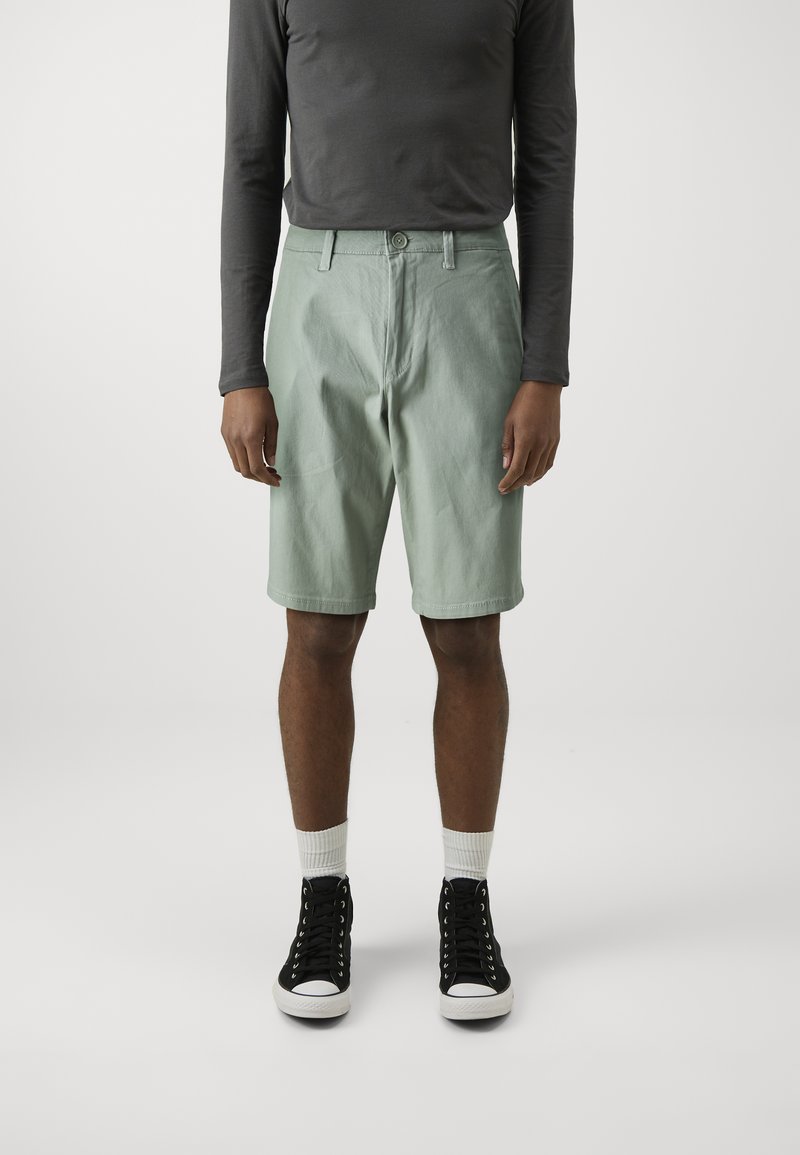 Pantalones cortos de color verde claro hechos de algodón, con un diseño clásico y cierre de botón, combinados con una camisa de manga larga oscura y zapatillas negras.