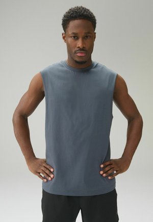 Pewny siebie mężczyzna stojący z rękami na biodrach, ubrany w szary bezrękawnik i czarne spodnie na jednolitym jasnym tle.