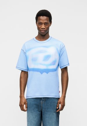 Mladý muž v bledomodrom voľnom tričku s vyblednutou bielou grafikou na prednej strane a modrých džínsoch, stojaci pred neutrálnym pozadím.