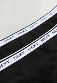 Musta kangas leveällä valkoisella vyötärönauhalla, jossa on toistuva "NEXT"-teksti mustana. Suloinen rakenne ilman lisäkuvioita tai koristeita.