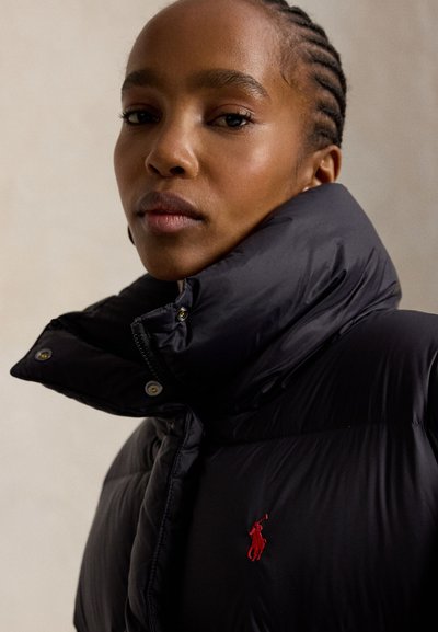 Kvinde med flettet hår iført en sort dunjakke med høj krave og et lille rødt broderet logo på brystet.