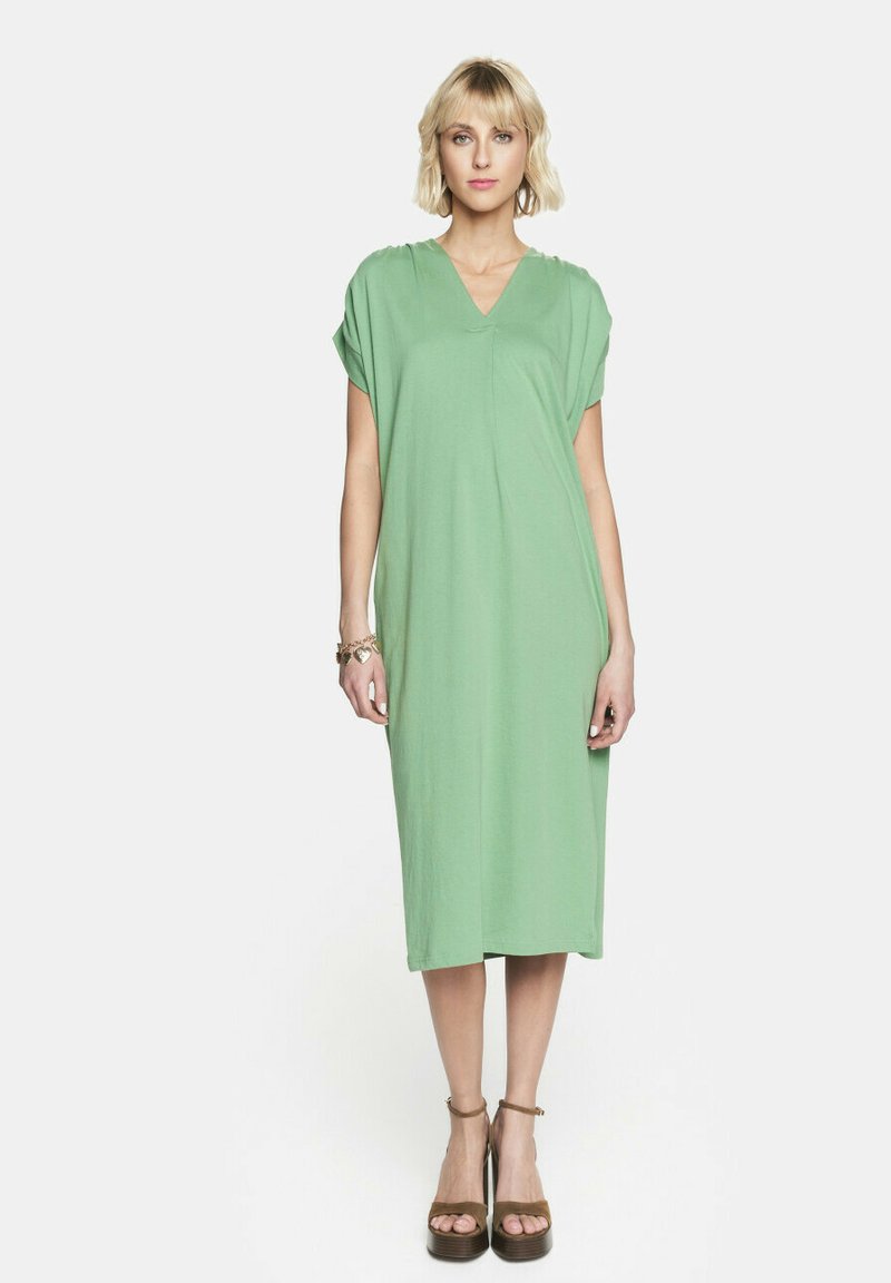 Jasnozielona sukienka o krótkim rękawie i dekolcie w serek, o luźnym kroju, długości do kolan, z gładkiego materiału. Modelka nosi sandały na platformie.