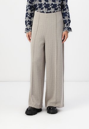 Pantalons larges en tissu gris clair texturé avec un motif en chevron. Présente des plis et un design taille haute. Associés à des chaussures noires imposantes.