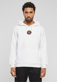 Hvit hettegenser laget av mykt stoff, med en frontlomme og en rund svart lapp med rød skrift. Ribbestrikkede ermer og nederkant.