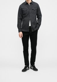 Camicia nera con bottoni e due tasche sul petto, indossata sopra una t-shirt bianca. Pantaloni aderenti neri e scarpe nere con dettagli bianchi.