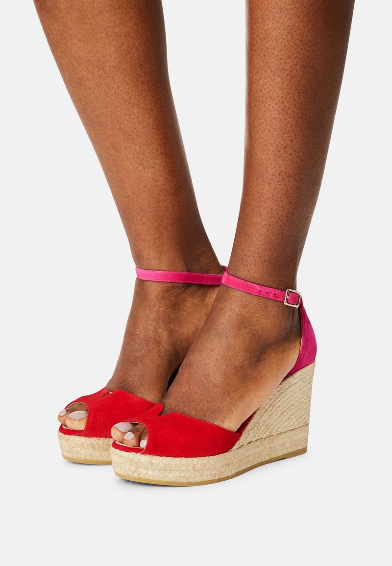 Pieds portant des sandales compensées à bout ouvert rouge et rose avec des brides à la cheville et des talons plateforme tissés beige sur un fond uni.