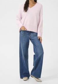 Pull en V rose clair, oversize, avec ourlet côtelé. Associé à un jean en denim bleu foncé ample et taille haute et des chaussures plates beiges.