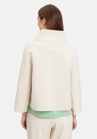 Giacca beige chiaro con colletto alto, maniche ampie e tessuto texturizzato. Indossata sopra una blusa verde menta con orlo arrotondato.