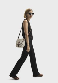 Small crossbody bag with beige fabric and brown leather accents, worn over a black sleeveless top and black trousers, paired with brown sandals.