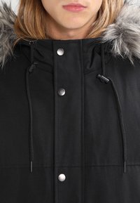 Svart parkas med huva i konstpäls, utrustad med dragskojusteringar, silverfärgade snap-knappar och en slät yta.