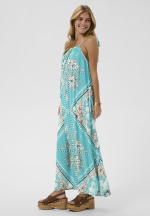 Mlada žena koja se smiješi, nosi plavu maksi haljinu bez rukava s cvjetnim uzorkom i smeđe sandale na platformu, uz zlatnu narukvicu manžetnu.