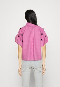 Blusa rosa con texture e maniche a sbuffo, con dettagli floreali neri sulle spalle e una silhouette aderente.