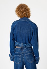 Chaqueta de mezclilla con un corte cropped, color azul, mangas largas, detalles con hebillas en los puños y la cintura, y un cuello clásico.