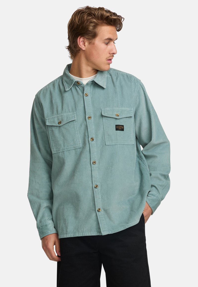Lys teal kordtrøje med to brystlommer, knaplukning og subtil ribbet tekstur. Har et logo-patch på den venstre lomme.