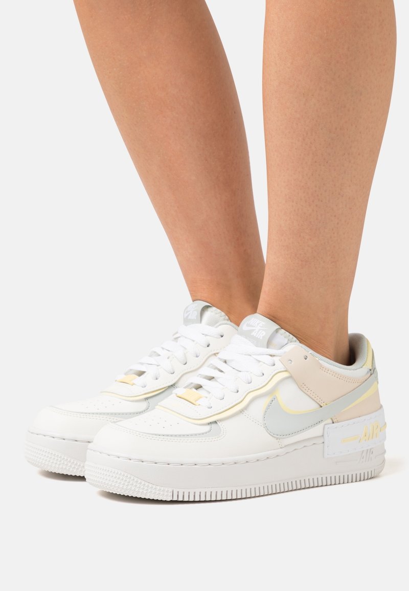 Paire de baskets blanches Nike Air avec des touches gris clair et beige portées aux pieds sur un fond clair uni.