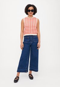 Débardeur tricoté sans manches avec un motif ondulé rouge et crème, associé à un pantalon en denim à jambes larges. Chaussures à talons blocs noires et lunettes de soleil surdimensionnées.