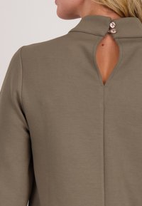 Vrouw in een olijfgroene top met een hoge kraag, een sleutelgatopening aan de achterkant en twee kleine knopen om de kraag vast te maken.