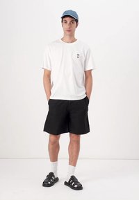 Hvit t-skjorte med korte ermer og liten grafikk, svarte shorts, hvite sokker og svarte sandaler. Blå caps fullfører antrekket.