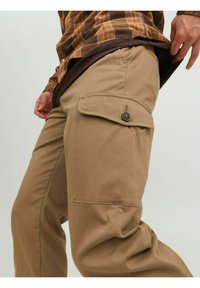 Pantaloni cargo beige con tasche laterali, dettagli con bottoni e un tessuto liscio e resistente. Presentano una vestibilità comoda e una leggera arricciatura alle ginocchia.