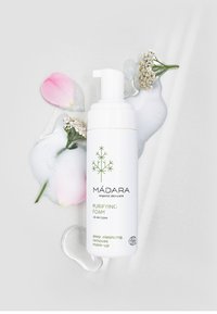 White pump bottle labeled "MÁDARA Purifying Foam" with green text. Surrounded by petals and soap bubbles on a light surface. 400 ml.