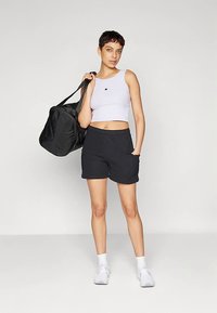 Ungdom som står i treningsklær: hvit kort topp, svarte shorts, hvite sneakers, og bærer en stor svart treningsbag over den ene skulderen.