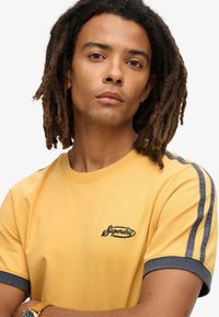 Geel katoenen T-shirt met korte mouwen, met blauwe accenten op de mouwen en een zwart logo geborduurd op de borst.