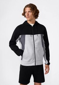 Grijs-zwart zip-up hoodie met raglanmouwen; voorzien van kleurvlakkenontwerp, verstelbare capuchon en zachte stoftextuur. Gecombineerd met zwarte shorts.
