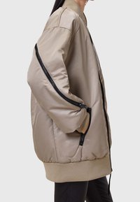 Beige Bomberjacke mit glattem, glänzendem Finish, schwarzen Reißverschlüssen, gerippten Bündchen und lockerem Schnitt.