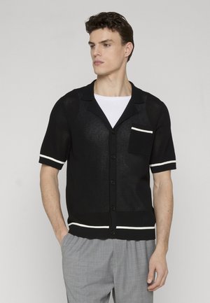 Uomo che indossa un cardigan nero a maniche corte in maglia con rifiniture bianche e tasca sopra una camicia bianca, abbinato a pantaloni grigio chiaro, con le mani nelle tasche.