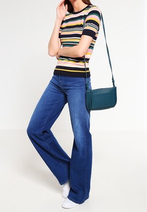 Donna con maglietta a maniche corte a righe, jeans blu a gamba larga, scarpe da ginnastica bianche e borsa a tracolla turchese, in piedi con una gamba incrociata.