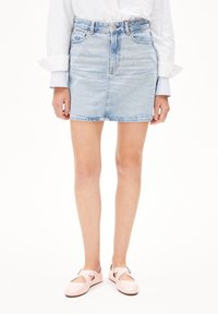 Jupe en denim bleu clair avec un design ajusté, présentant une taille haute, un ourlet brut et des poches avant. Portée avec des ballerines roses brillantes.