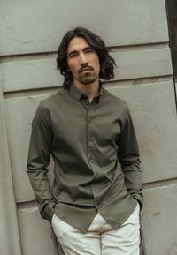 Mann mit mittellangen dunklen Haaren und Bart, der ein olivgrünes Hemd mit Knöpfen und weiße Hose trägt und an einer strukturierten Wand lehnt.