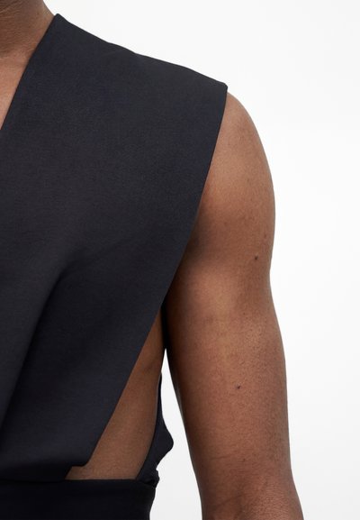 Κοντινό πλάνο του άνω βραχίονα και του ώμου ενός ατόμου που φοράει ένα μαύρο αμάνικο ένδυμα με ευρεία κάλυψη ώμων και πλευρικές ανοίγματα.