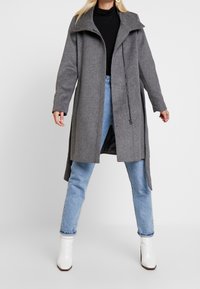 Cappotto grigio in lana con un colletto alto, caratterizzato da un orlo asimmetrico e un dettaglio con zip. Accoppiato con jeans azzurri chiari e stivaletti bianchi alla caviglia.