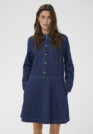 Woman wearing a long-sleeve dark blue denim dress with front buttons, chest pocket, collar, and hands in side pockets.