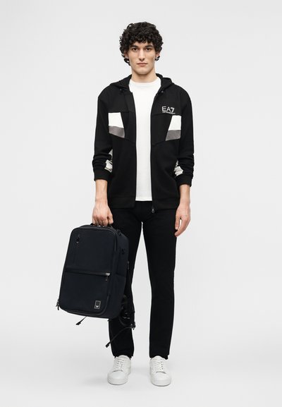 Νέος άνδρας με ασπρόμαυρο μπουφάν και παντελόνι κρατάει μαύρο σακίδιο πλάτης με λευκά αθλητικά παπούτσια, πάνω σε απλό λευκό φόντο.